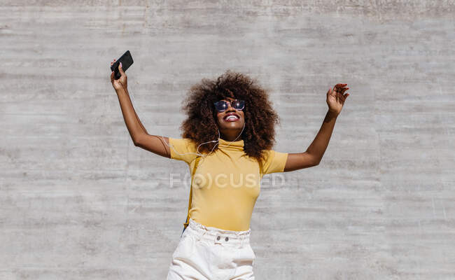 Чорна жінка з афро волоссям слухає музику на мобільному телефоні перед сірою стіною — стокове фото