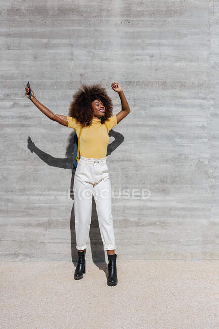 Black woman with afro hair listening to music on mobile in front of a gray wall — Fotografia de Stock