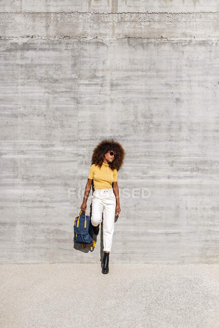 Schwarze Frau mit Afro-Haaren hört Musik auf dem Handy vor einer grauen Wand und hält einen Rucksack mit der Hand — Stockfoto