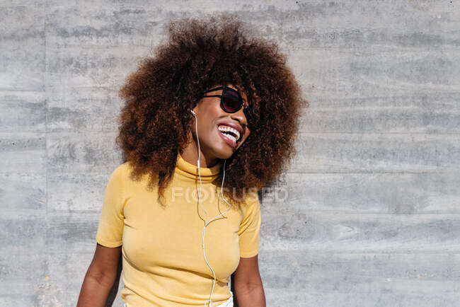 Black woman with afro hair listening to music on mobile in front of a gray wall — Photo de stock