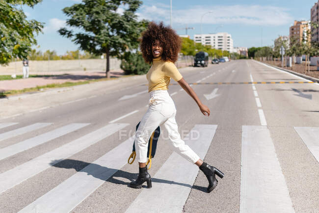 Черная женщина с афроволосами переходит улицу с рюкзаком в руке — стоковое фото