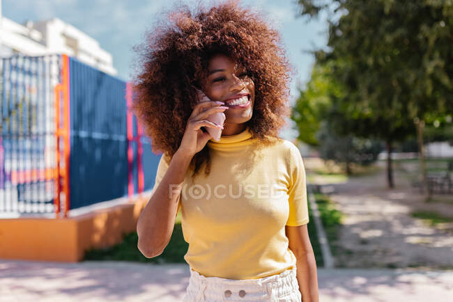 Mujer negra con pelo afro hablando por teléfono mientras camina por la calle - foto de stock
