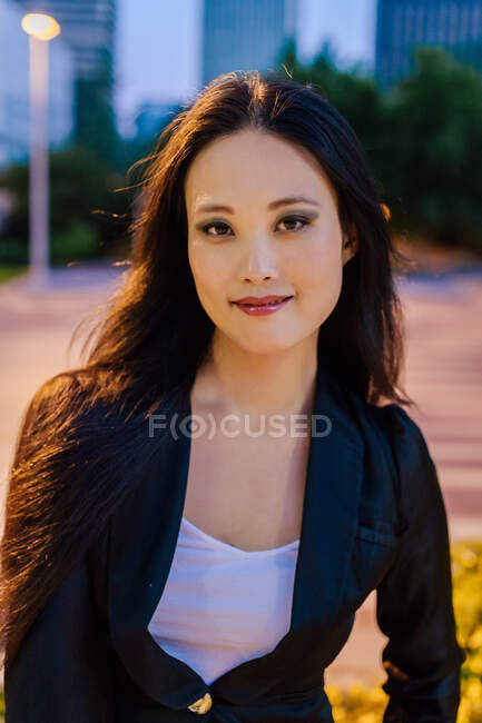 Cheerful Asian female entrepreneur in smart casual outfit looking at camera on background of city in evening — Stock Photo