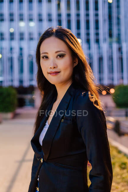 Asian businesswoman in elegant outfit looking at camera while standing on street in downtown on background during evening — Stock Photo