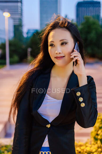 Entzückte asiatische Unternehmerin steht abends in der Straße von Megapolis und telefoniert — Stockfoto