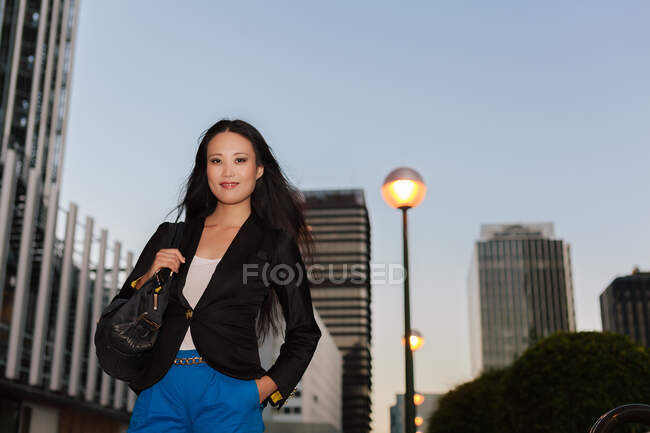 Bajo ángulo de la mujer de negocios asiática en traje elegante mirando a la cámara mientras está de pie con la mano en el bolsillo en la calle en el centro de la ciudad en el fondo del cielo puesta del sol - foto de stock