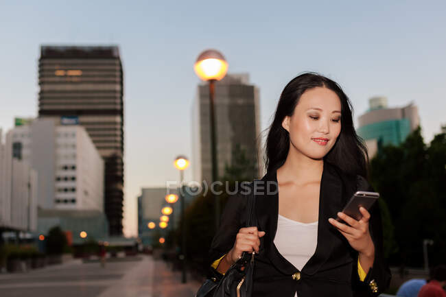 Delighted Asian female entrepreneur standing in street of megapolis in evening and messaging on mobile phone — Stock Photo
