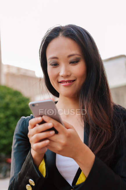 Счастливая азиатская предпринимательница в смарт-случайном стиле стоит на улице в центре города просматривая на мобильном телефоне — стоковое фото