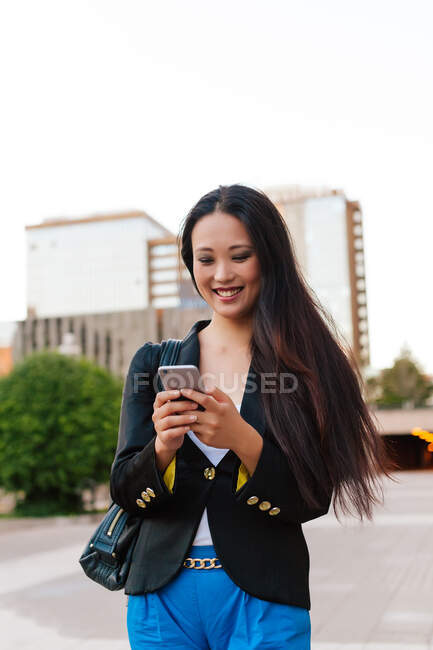 Heureux entrepreneur asiatique femme dans le style décontracté intelligent debout dans la rue du centre-ville de navigation sur téléphone mobile — Photo de stock