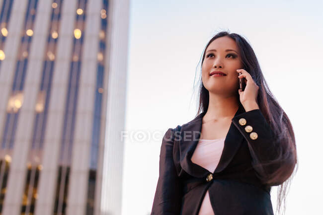 Von unten steht eine asiatische Unternehmerin in schickem, lässigem Stil in der Innenstadt und telefoniert mit dem Handy, während sie wegschaut — Stockfoto