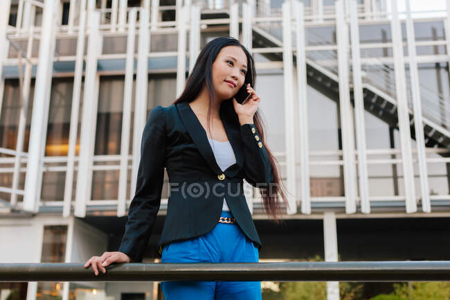 Desde abajo de la mujer empresaria asiática en el estilo casual inteligente de pie en la calle del centro de la ciudad y hablando por teléfono móvil mientras mira hacia otro lado - foto de stock