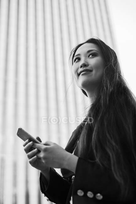 From below of happy Asian female entrepreneur in smart casual style standing in street of downtown browsing on mobile phone while looking away — Stock Photo