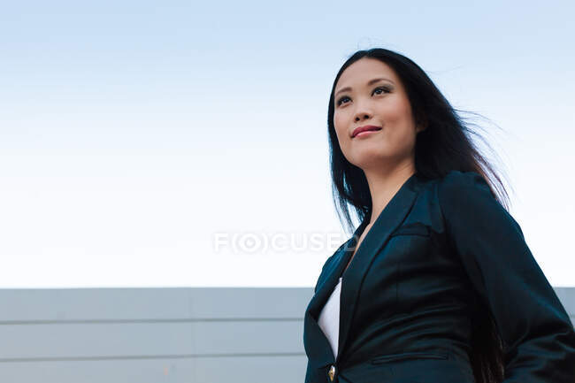 Снизу уверенная в себе азиатская предпринимательница, идущая по улице рядом с городским зданием и смотрящая в сторону — стоковое фото