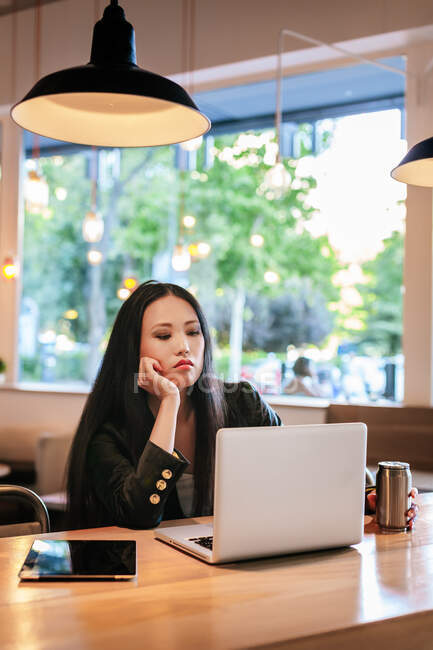 Empresária asiática exausta sentada à mesa com bebida energética em lata de alumínio e lendo informações sobre laptop enquanto trabalhava remotamente no café — Fotografia de Stock