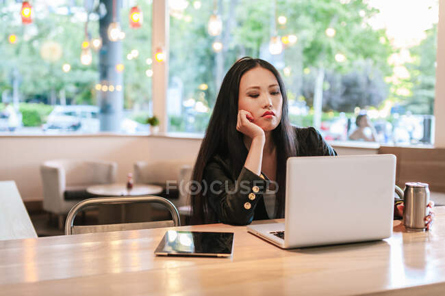 Уставшая азиатская предпринимательница сидит за столом с энергетическим напитком в алюминиевой банке и читает информацию на ноутбуке, работая дистанционно из кафе — стоковое фото