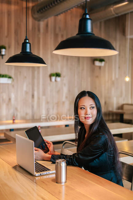 Contenu entrepreneur ethnique féminin assis à table avec tablette et ordinateur portable tout en travaillant sur le projet et en regardant ailleurs — Photo de stock