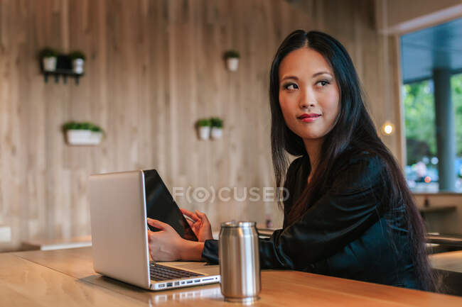 Conteúdo empresária étnica sentada à mesa com tablet e laptop enquanto trabalhava no projeto e desviava o olhar — Fotografia de Stock