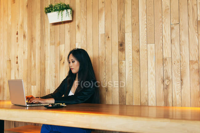Femme entrepreneur asiatique concentrée assise à table dans un café et tapant sur le netbook tout en travaillant sur un projet en ligne à distance — Photo de stock