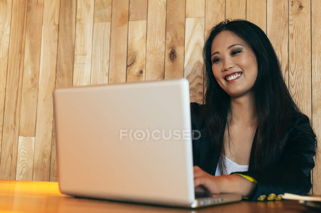 Positiva mujer empresaria asiática sentada en la mesa en la cafetería y escribiendo en netbook mientras sonríe y trabaja en el proyecto en línea de forma remota - foto de stock
