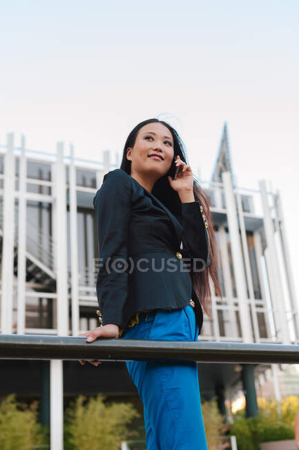 From below side view of Asian female entrepreneur in smart casual style standing in street of downtown and talking on mobile phone while looking away — Stock Photo