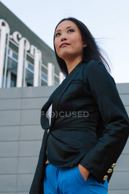 Baixo ângulo de determinado bem vestido empresária asiática em pé com a mão no bolso na rua da cidade e olhando para longe — Fotografia de Stock