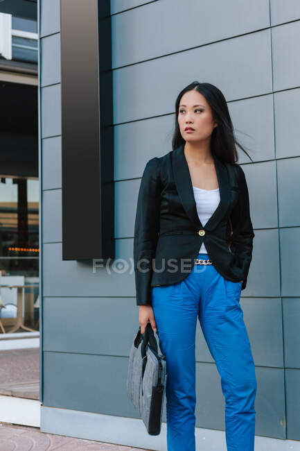 Самоуверенная азиатская предпринимательница, идущая по улице рядом с городским зданием и отворачивающаяся — стоковое фото