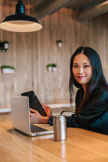 Contenida empresaria étnica sentada a la mesa con tablet y laptop mientras trabaja en el proyecto y mira hacia otro lado - foto de stock