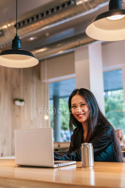 Conteúdo empresária étnica sentada à mesa com laptop trabalhando no projeto e desviando o olhar — Fotografia de Stock