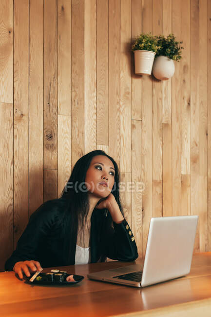 Занятая азиатская предпринимательница, сидящая за столом в кафе во время еды суши и работы над удаленным проектом с помощью нетбука — стоковое фото