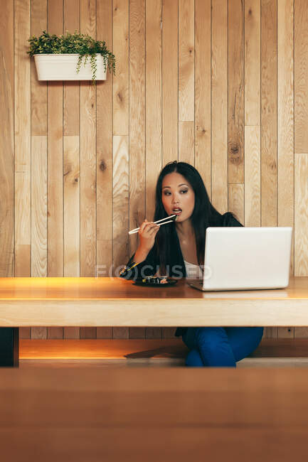 Empresaria asiática ocupada sentada a la mesa en la cafetería mientras come sushi y trabaja en un proyecto remoto a través de netbook - foto de stock