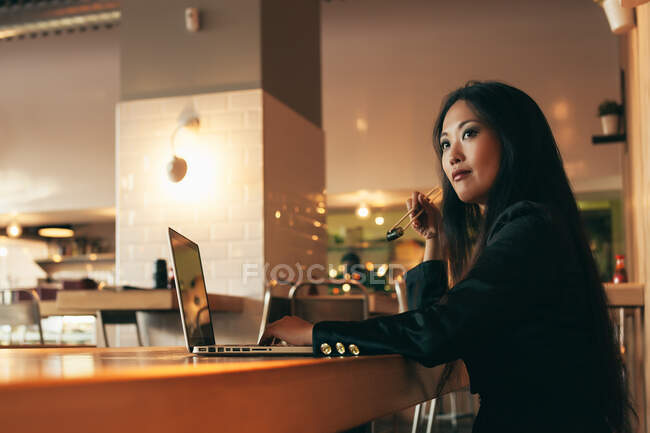 Side view of busy Asian female entrepreneur sitting at table in cafe while eating sushi and working on remote project via netbook — Stock Photo
