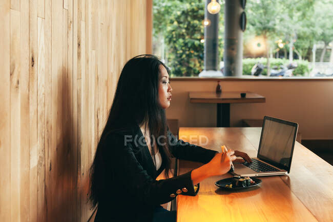 Vista lateral de la ocupada empresaria asiática sentada en la mesa en la cafetería mientras come sushi y trabaja en un proyecto remoto a través de netbook - foto de stock