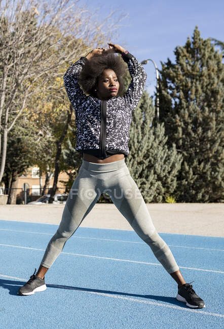 Fit African American female athlete standing at stadium and stretching body while warming up with raised arms before workout on sunny day and looking away — Stock Photo