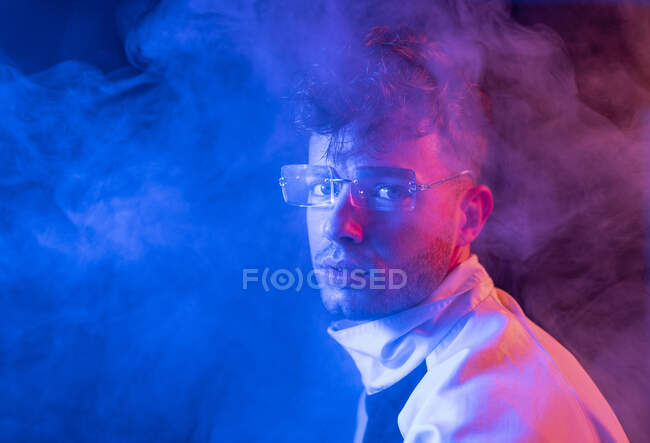 Vista lateral de homem jovem pensativo em roupa elegante e óculos olhando para a câmera enquanto estava em pé no quarto neon fumegante escuro — Fotografia de Stock