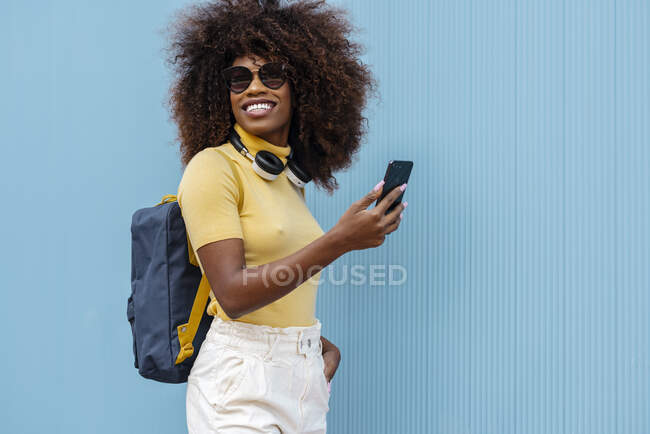 Glad ethnic female with Afro hairstyle and headphones taking self portrait on cellphone on blue background — Stock Photo