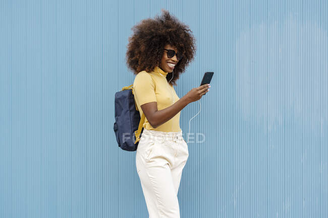 Радісна етнічна жінка з африканською зачіскою та навушниками, які дивляться телефон на синьому тлі. — стокове фото