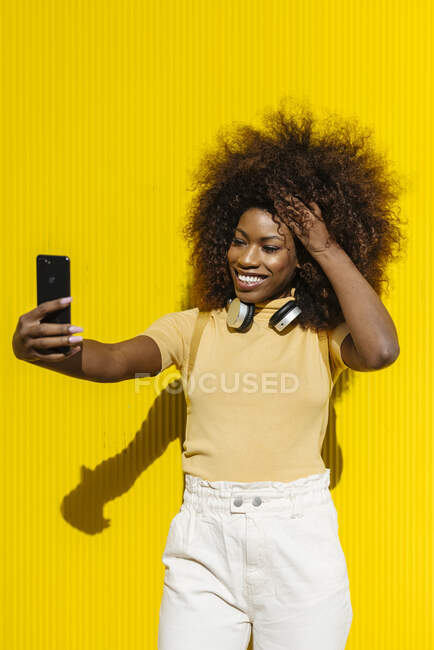 Glad ethnic female with Afro hairstyle and headphones taking self portrait on cellphone on yellow background with shadow outdoors — Fotografia de Stock