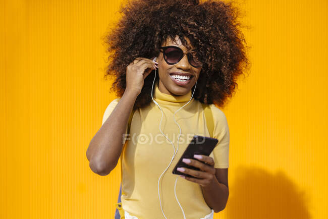 Content young ethnic female in sunglasses with Afro hairstyle browsing internet on cellphone while listening to music on yellow background — Fotografia de Stock