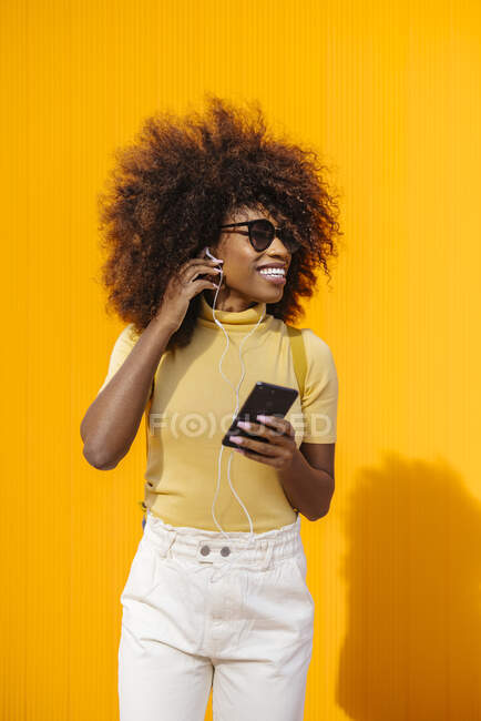 Содержание молодых этнических женщин в солнцезащитных очках с прической Afro, просматривающих интернет по сотовому телефону, слушая музыку на желтом фоне — стоковое фото
