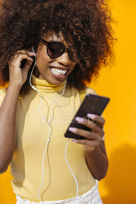 Зміст молодої етнічної жінки в сонцезахисних окулярах з африканською зачіскою переглядає інтернет на мобільному телефоні, слухаючи музику на жовтому тлі. — стокове фото