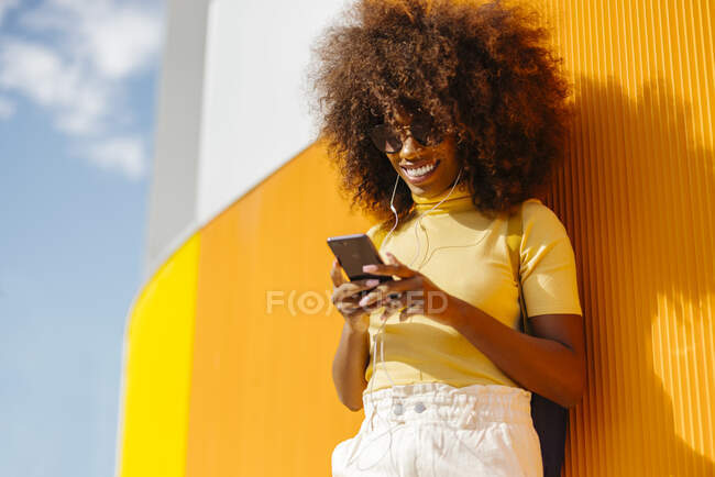 Contenuto giovane femmina etnica in occhiali da sole con acconciatura afro navigare in internet sul cellulare durante l'ascolto di musica su sfondo giallo — Foto stock