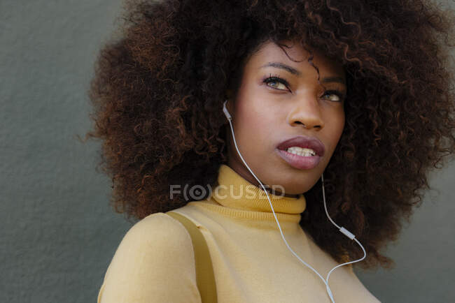 Giovane contemplativa etnica femminile con acconciatura afro ascoltare la canzone dagli auricolari mentre distoglie lo sguardo su sfondo grigio — Foto stock