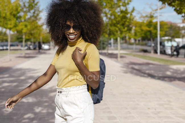 Cheerful young African American female traveler with rucksack standing on walkway in city on sunny day — Stock Photo