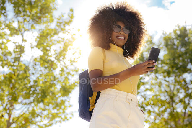 From below of glad ethnic female in earphones and sunglasses taking selfie on cellphone under blue sky in sunlight — Fotografia de Stock