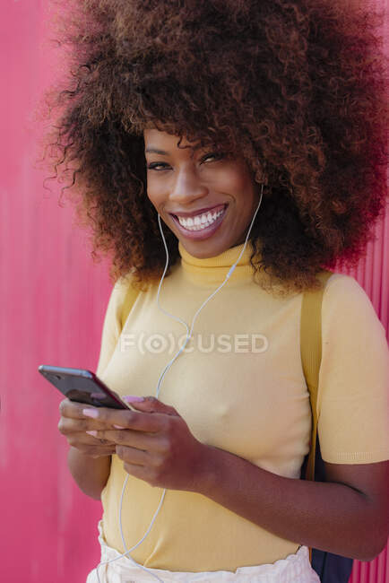 Zufriedene junge ethnische Frau mit Afro-Frisur surft im Internet auf dem Handy, während sie Musik auf rosa Hintergrund hört — Stockfoto