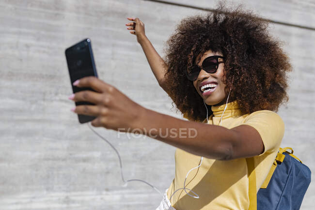 Feliz mujer étnica en auriculares y gafas de sol tomando selfie en el teléfono celular cerca de la pared gris a la luz del sol - foto de stock