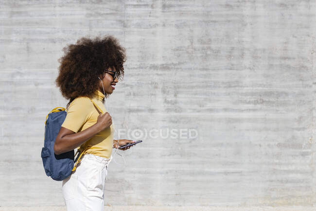 Glad ethnic female with Afro hairstyle and headphones walking near grey wall — Photo de stock