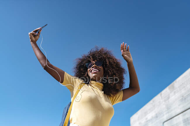 Von unten eine fröhliche ethnische Frau mit Kopfhörern und Sonnenbrille, die unter blauem Himmel im Sonnenlicht ein Handy-Selfie macht — Stockfoto