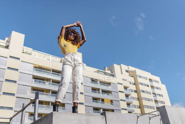 From below of cheerful African American female in casual clothes and sunglasses standing with raised arms under blue cloudy sky — Fotografia de Stock