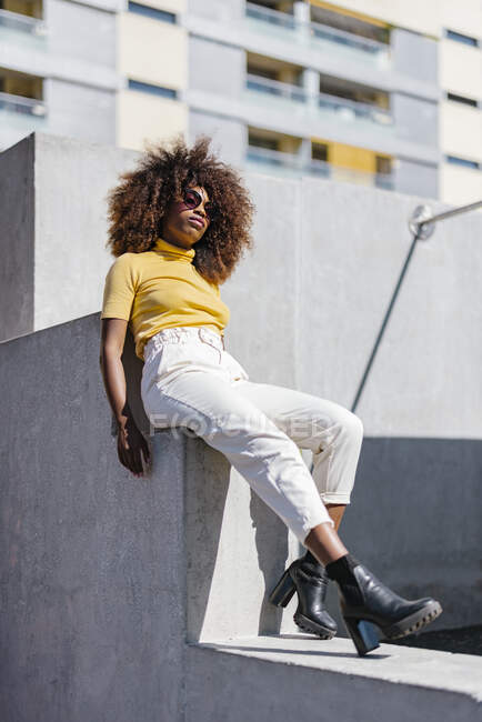 From below of African American female in casual clothes and sunglasses sitting on facade on street — Stock Photo